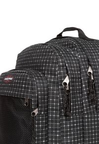 Mochila de tela negra con patrón de cuadrícula blanca, que cuenta con dos compartimentos con cremallera y tiradores metálicos. Se ve un bolsillo lateral texturizado.
