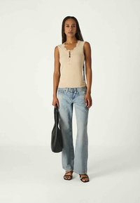 Camisola de alça em algodão bege com acabamento de renda, placket de três botões, combinada com jeans justos azuis claros; modelo segura uma bolsa de ombro preta.