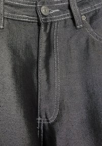 Jean en denim noir à texture lisse, présentant des coutures blanches contrastantes, un bouton en métal et une braguette à fermeture éclair.