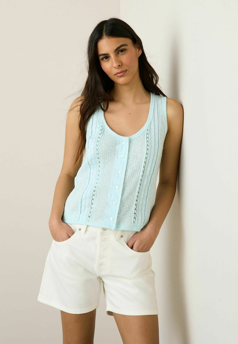 Žena s dlhými tmavými vlasmi, oblečená v svetlomodrom bezrukávovom zapínacom topánku a bielych šortkách, stojaca s rukami v vreckách opretá o stenu.