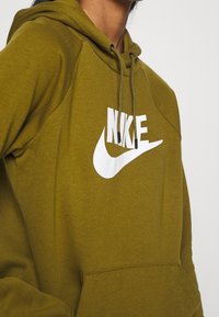 Olivově zelená mikina s kapucí z měkké látky, která má b�ílé logo Nike na hrudi. Obsahuje stahovací kapuci a klokaní kapsu.