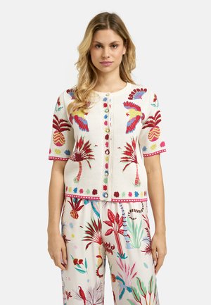Woman wearing a white short-sleeve buttoned top and pants with colorful tropical bird and plant patterns.