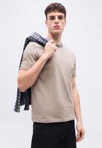 Νεαρός άνδρας που φοράει μπεζ μπλουζάκι και σκούρο τζιν κρατάει πουκάμισο με μπλε και γκρι καρό πάνω στον ώμο σε απλό φόντο.