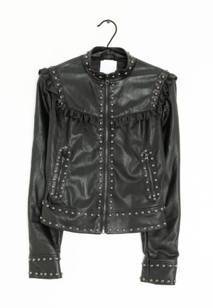 Veste en cuir noire avec fermeture éclair, ornements cloutés, détails à volants sur les épaules et poches zippées, suspendue sur un cintre noir.