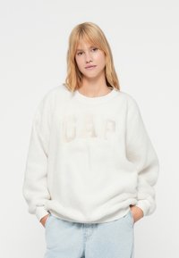 Hvit fleecegenser med rund hals og lange ermer. Har "GAP"-logo i subtil glans. Kombinert med lyseblå jeans.