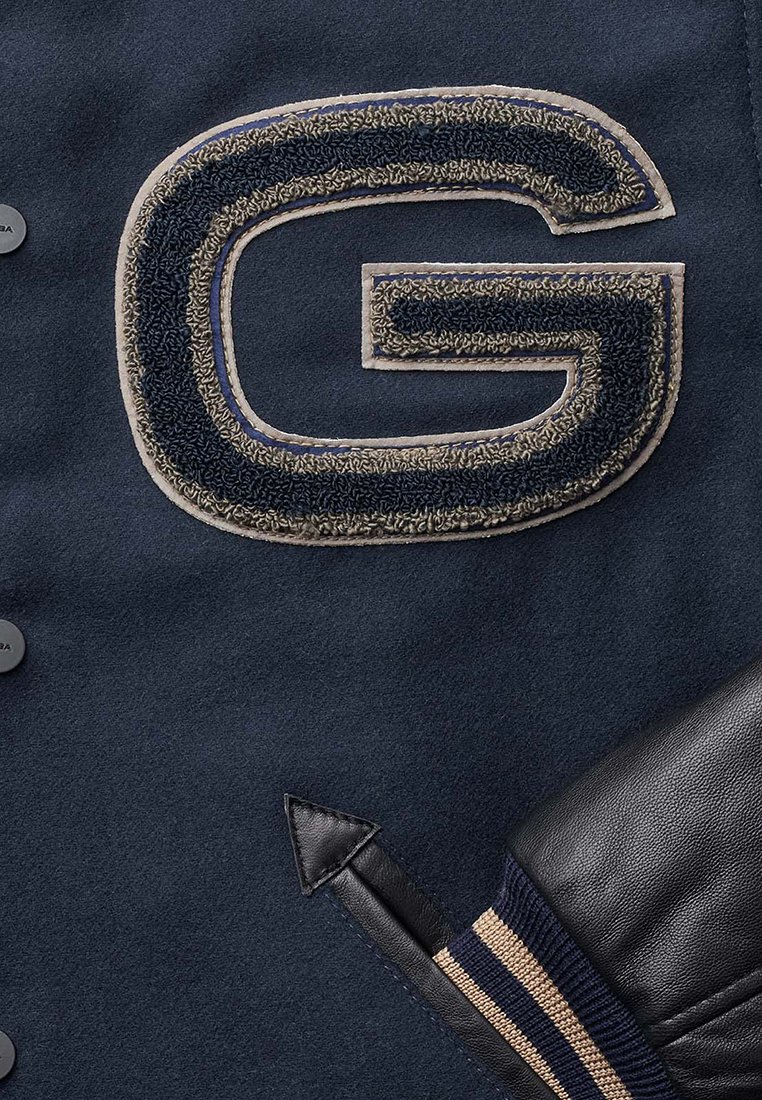 Marineblauwe wollen jas met een grote gestructureerde "G"-patch in donkerblauw en beige, met zwarte leren accenten en gestreepte ribkousen.