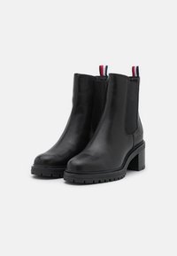 Botas de cuero negro hasta el tobillo con paneles elásticos laterales, punta redonda y tacón grueso. Presenta una lengüeta de tiro en colores rojo, blanco y azul.