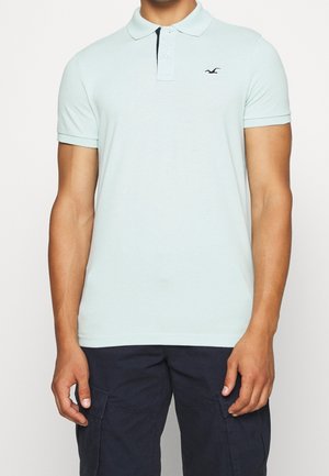 Hombre que lleva una camisa polo de manga corta azul claro con un pequeño logotipo de un pájaro negro en el pecho izquierdo, combinada con pantalones cargo oscuros.