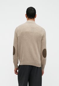 Bežs adīts džemperis ar garām piedurknēm, kas izceļas ar brūnām elkoņu plecu uzlikām un ribotām aprocēm. Tīrs, vienkāršs dizains ar brīvu piegriezumu.