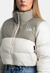Giacca trapuntata in beige e bianco, con colletto alto, zip frontale e logo sul petto. Tessuto testurizzato con sezioni cucite.