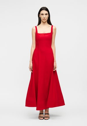 Γυναίκα με μακριά σκούρα μαλλιά, που φοράει ένα κόκκινο φόρεμα μέχρι το πάτωμα χωρίς μανίκια, με τετράγωνη λαιμόκοψη και μαύρα ανοιχτά παπούτσια με τακούνι, στέκεται μπροστά σε ένα απλό φόντο.