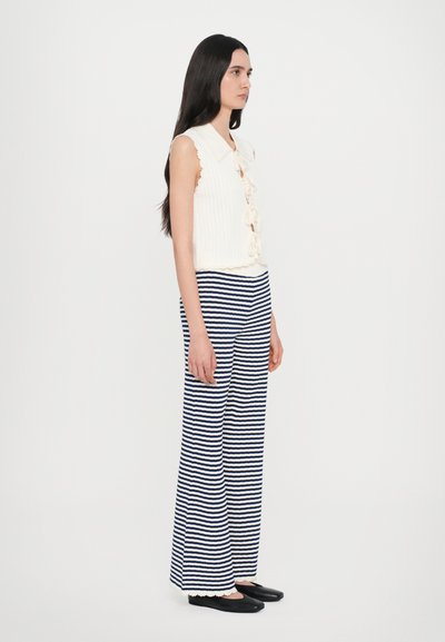 Haut sans manches blanc à côtes avec un col et des nœuds décoratifs, assorti à un pantalon à jambes larges rayé bleu marine et blanc et des chaussures plates noires.