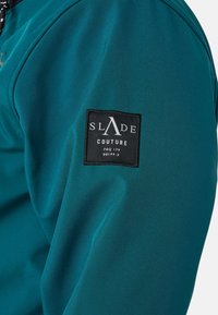 Giacca teal con tessuto liscio e strutturato. Presenta una toppa quadrata nera con il logo "SLADE COUTURE" sul maniche sinistro. Dettaglio con zip visibile.