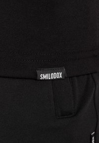 Material textil negro con un acabado suave, que presenta una pequeña etiqueta blanca que dice "SMILODOX". Costuras visibles a lo largo de los bordes.