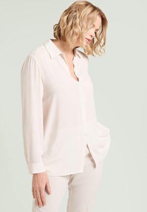 Mujer con cabello rubio ondulado, vestida con una blusa crema de manga larga con botones y pantalones a juego, de pie frente a un fondo claro.