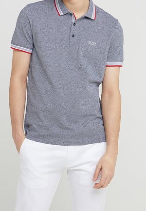 Man draagt een kort grijs poloshirt met zwart, wit en rood randje en witte broek, handen gedeeltelijk zichtbaar.