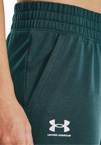 Dunkelgrüne Sporthosen mit einem elastischen Bund, seitlichen Taschen und einem weißen Under Armour-Logo auf der unteren Seite.