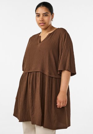 Vrouw met donker haar in een staart gebonden, die een losse, gestructureerde bruine jurk draagt over een witte broek, staand voor een effen witte achtergrond.