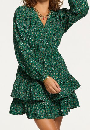 Femme portant une robe mini à manches longues verte à fleurs avec taille froncée, collier en or et bracelet en or, posant avec un bras levé.