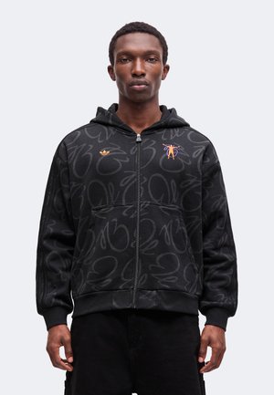 Jeune homme portant un sweat à capuche noir zippé avec un motif abstrait, un logo orange sur la poitrine et un pantalon noir, debout devant un fond gris.