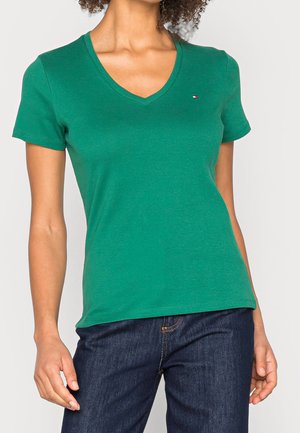 Vrouw draagt een groen T-shirt met V-hals en korte mouwen en donkerblauwe spijkerbroek, staand met één hand ontspannen naast zich.