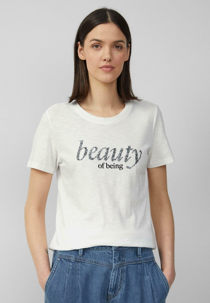 T-shirt en coton blanc à manches courtes, avec un design de texte noir et argent qui dit "la beauté d'être". Coupe décontractée, col ras du cou.