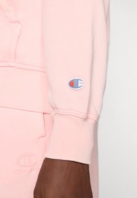 Sweatshirt roz deschis cu manșete striate, având un mic logo brodat în albastru și roșu pe mânecă și detalii de cusătură subtile.