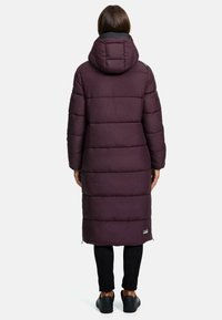 Dolga vijolična puffer jakna s kapuco, ki ima vodoravno šivanje in logotip na robu. Nošena je preko črnih hlač in čevljev.