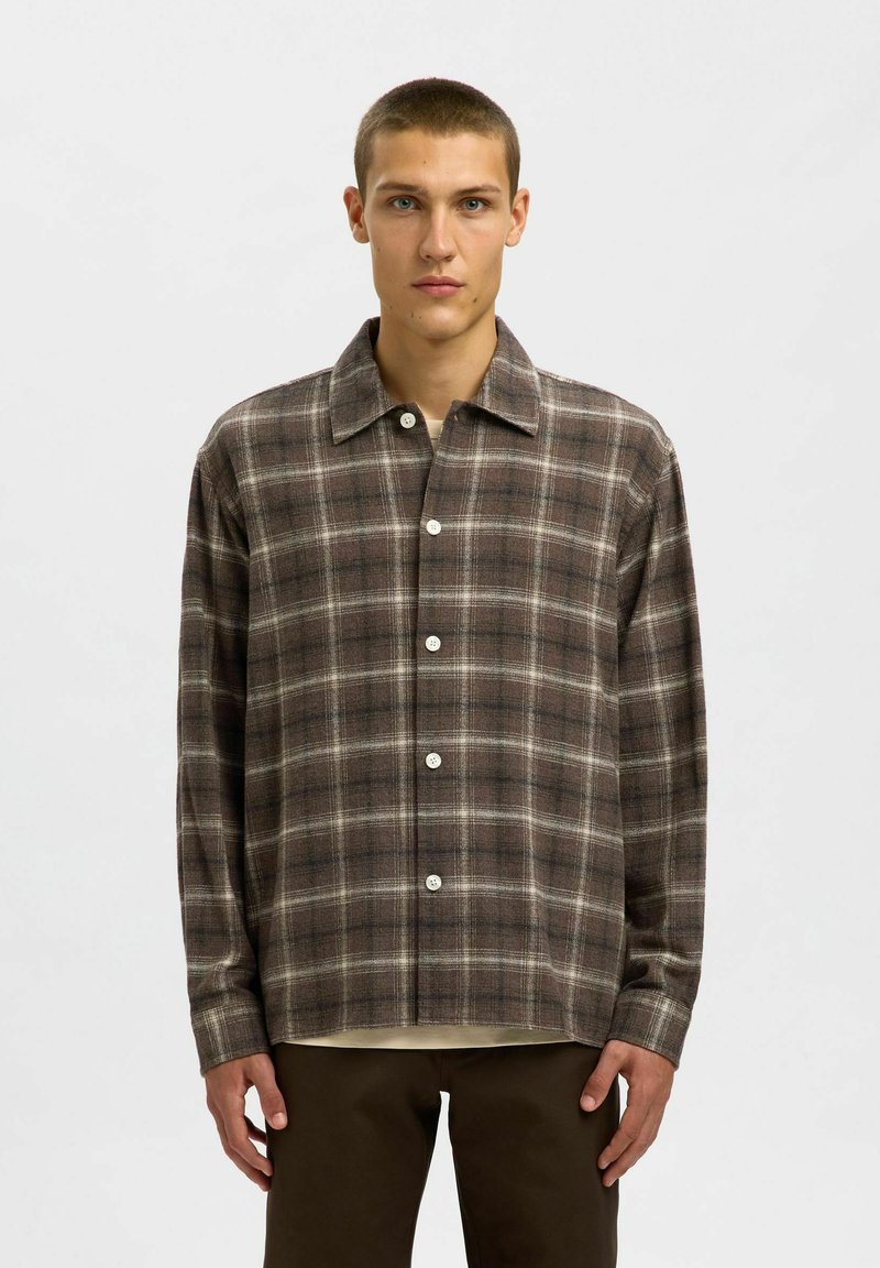 Jaunais vīrietis, kurš valkā pogātu brūnu platas svītras krekliņu uz beigtas apakšveļas, kombinējot to ar tumši brūnām biksēm, stāv uz vienkārša fona.