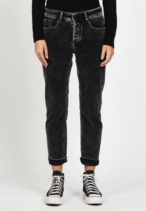 Person, die schwarze, verwaschene Jeans mit umgeschlagenen Säumen an den Knöcheln und schwarze High-Top-Sneaker mit weißen Schnürsenkeln trägt, steht vor einem einfarbigen Hintergrund.