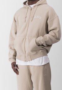 Sudadera con cremallera en color beige, hecha de tela suave, con un bolsillo canguro, puños acanalados y detalles de logo bordados en el pecho y la manga.