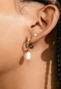 Guld hoop ørering med en hængende hvid perle og en mindre hvid perle som sten. Glat overflade, blank finish og subtile detaljer på hoop'en.