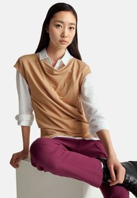 Béžový přehozený top přes bílou košilí s límečkem, doplněný burgundskými kalhotami a černými šněrovacími botami. Hladká látka, moderní design.