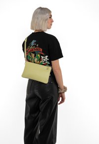 Pale green crossbody bag with a smooth texture, rectangular shape, and zipper closure, worn over a black graphic t-shirt and leather pants.