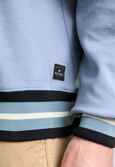 Jasnoniebieski sweter z teksturowanego materiału z czarną kwadratową metką z napisem "PS Paul Smith". Rękawy ozdobione są paskami w odcieniach niebieskiego, jasnoniebieskiego i czarnego.
