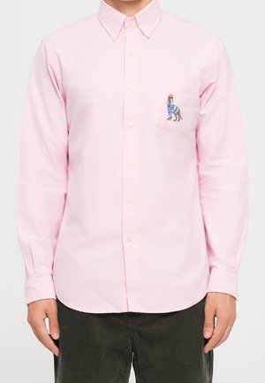 Man draagt lichtroze overhemd met knopen, met een geborduurde hond in een blauwe jas en rood hoedje op het linkerborstzakje.