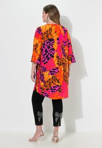 Bunte Tunika mit einem lebhaften, orange, pink und schwarzen abstrakten Muster, kombiniert mit schwarzen Hosen, die mit mit Juwelen besetzten Schleifenakzenten am Saum versehen sind.