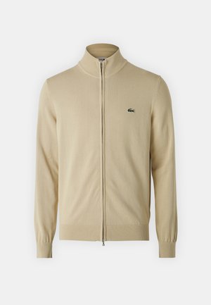 Beige zip-up kofta med hög krage och ribbade ärmslut. Broderad grön logotyp på bröstet. Mjuk stickad material, strömlinjeformat design.