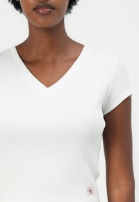 White ribbed short-sleeve top featuring a V-neck and a small CK logo patch on the lower side, with a fitted silhouette and smooth texture.