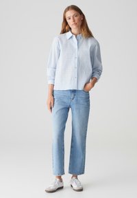 Camicia button-up a righe blu chiaro con colletto, maniche lunghe e vestibilità rilassata, abbinata a jeans a gamba dritta in denim chiaro e sneakers bianche.