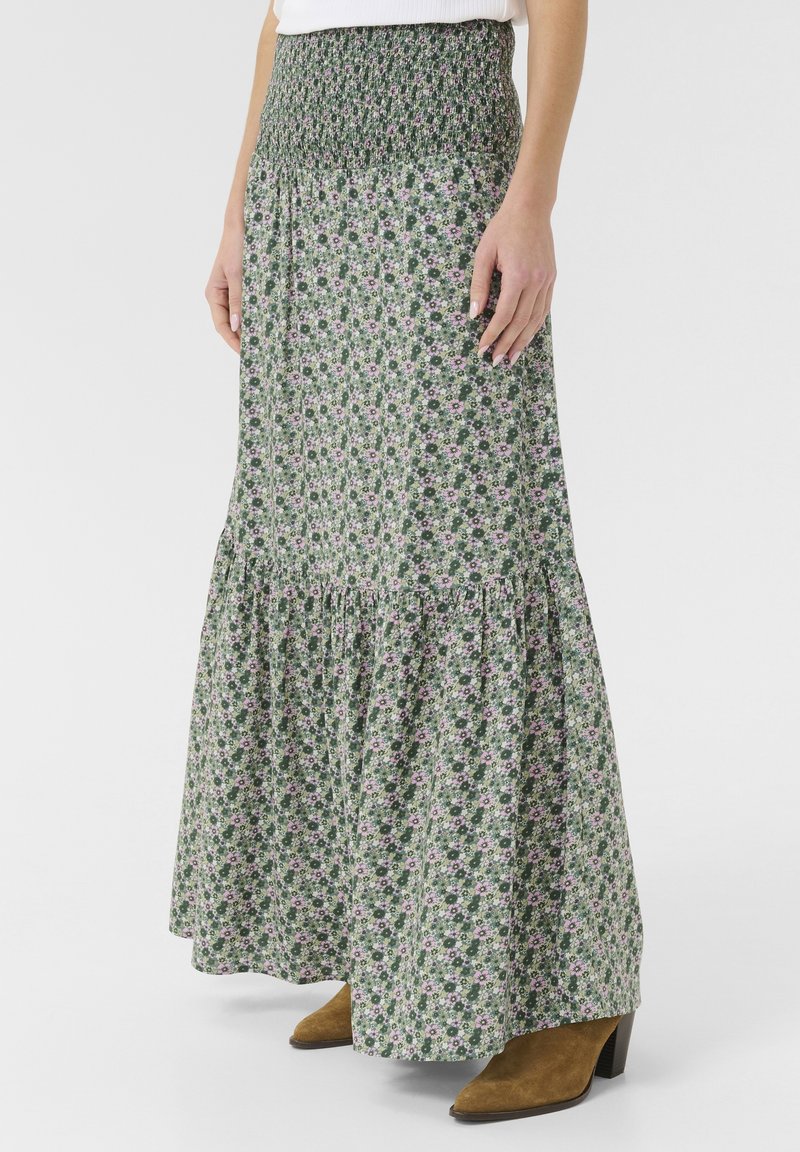 Blomstret maxi-nederdel med en smocket talje, prydet med grønne og pink mønstre, tiered kant og et flydende design. Brune hæle støvler synlige.
