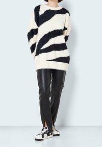 Przesadnie duży kremowy sweter z czarnymi abstrakcyjnymi paskami, zestawiony z dopasowanymi czarnymi spodniami z eko skóry oraz monochromatycznymi trampkami.