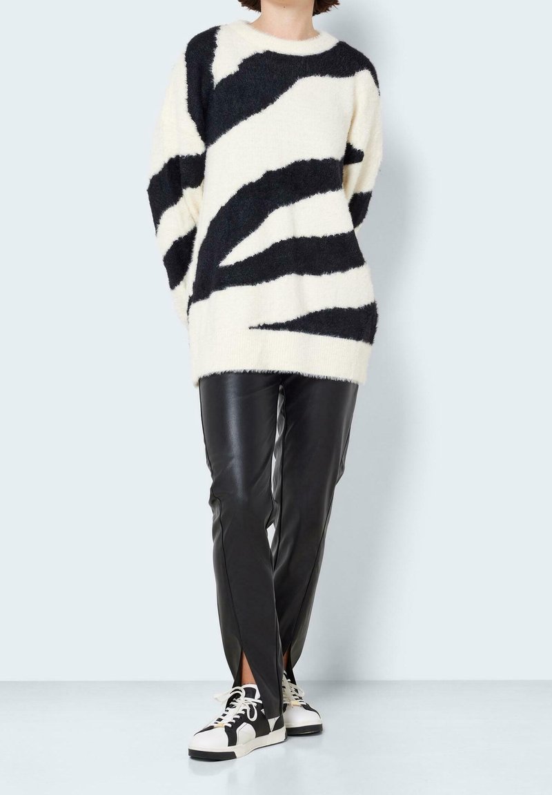 Przesadnie duży kremowy sweter z czarnymi abstrakcyjnymi paskami, zestawiony z dopasowanymi czarnymi spodniami z eko skóry oraz monochromatycznymi trampkami.