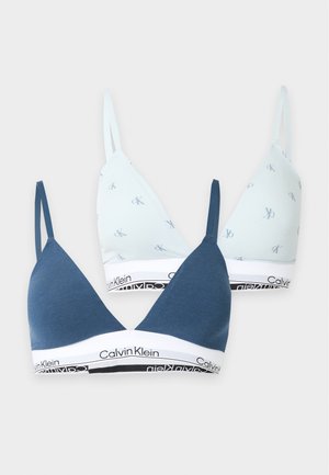 Žena s dlhými tmavými vlasmi, ktorá má na sebe biely Calvin Klein bralette a spodnú bielizeň, stojí bosá na jednoduchom svetlom pozadí.