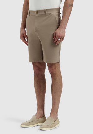 Man wearing beige tailored shorts, beige slip-on shoes, and a white shirt, standing with hands relaxed by his sides.