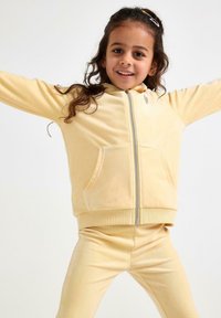 Gul velour træningsdragt med en lynlåshætte og forlommer. De tætsiddende bukser matcher toppen og har en glat tekstur samt ribbede manchetter.