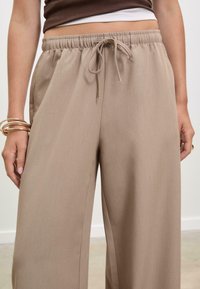 Beige wijde broek met een elastische tailleband en een trekkoorddetail. Glad materiaal met een lichte glans; getailleerde pasvorm langs de benen.