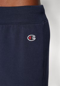 Nahaufnahme von marineblauen Sweatpants mit elastischem Bund und kleinem, nahe der Tasche gesticktem Champion-Logo.