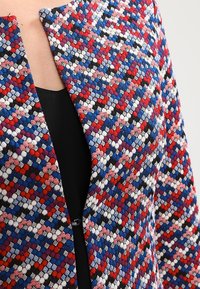 Lähellä kuvattu monivärinen, tekstuurinen takki, jossa on geometrinen kuosi, pukeutuneena mustan paidan päälle.