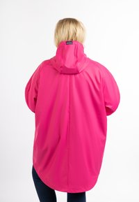 Personne aux cheveux blonds portant un imperméable rose vif avec une capuche, vue de dos sur fond blanc.